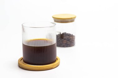 Bir bardak taze demlenmiş kahvenin yer aldığı minimalist natüralist bir imaj, yuvarlak bir ahşap bardak altlığında dinleniyor, yanında da el değmemiş bir kahve çekirdeğine karşı izole edilmiş, kavrulmuş kahve çekirdekleri dolu bir kavanoz var.