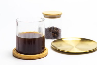 İyi aydınlatılmış bir stüdyo çekiminde, altın bir tepsi ve bir kavanoz kahve çekirdeğinin yanındaki küçük bir ahşap bardak üzerinde koyu kahve setiyle dolu temiz bir kaburgalı bardak sergilenir.