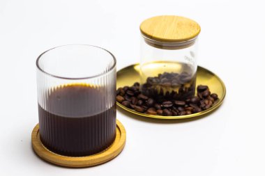 Minimalist ve zarif bir çekim, bir bardak koyu kahveyi ahşap bir altlığın üzerinde sergiliyor, yanında da el değmemiş beyaz bir zemin üzerinde bir kavanoz kahve çekirdeği var. Bu resim