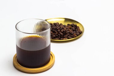 Bir bardak koyu kahvenin yer aldığı davetkar bir görüntü, ahşap bir altlığın üzerine kurulmuş, yanında da kavrulmuş kahve çekirdekleri ile dolu altın bir tepsi. Parlak renge karşı kompozisyon temiz ve modern.