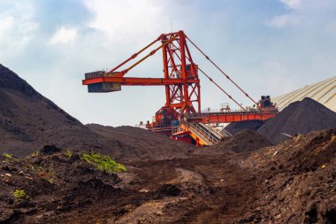 Görüntü bir kömür madeni ve ağır endüstriyel makinelerini gösteriyor. Site, operasyonun boyutunu ve kaynak çıkarma sürecini vurgulayan büyük kömür yığınlarıyla dolu. Şey...