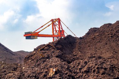 Madencilik ya da inşaat alanında ağır yük taşıyan bir vinci gösteren canlı bir görüntü. Parlak turuncu makine, koyu toprak ve mavi gökyüzüne karşı zıt yöndedir.