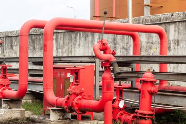 Beton bir zemine dayanan vanaları ve yangın musluğu olan endüstriyel kırmızı boru sisteminin ayrıntılı bir görüntüsü, altyapının karmaşıklığını ve endüstriyel süreçler için önemini gözler önüne seriyor.