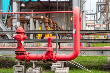 Görüntü, bir işleme tesisi veya fabrikanın arka planına kurulmuş bir dizi kırmızı boru ve vanayla birlikte bir yangın musluğu ve karmaşık endüstriyel boru hattı altyapısını gösteriyor. Fotoğraf şunu vurguluyor:
