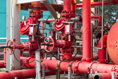 Yakın plan çekimde bir tesis içindeki karmaşık kırmızı endüstriyel boru hatları, vanalar ve göstergeler sergileniyor. Metalik yapı, gerekli karmaşık mühendislik ve bakımı vurguluyor