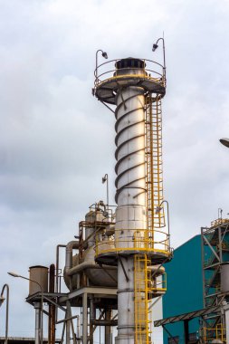 Bu görüntü, bulutlu bir gökyüzüne karşı duran uzun bacalı bir endüstriyel tesis altyapısını gösteriyor. Borular, tanklar ve metal yapılardan oluşan karmaşık ağ,