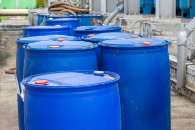 Bir endüstriyel depolama alanında kırmızı kapaklı büyük mavi plastik varillerden oluşan bir grup düzenleniyor. Görüntü, kimyasal veya atık maddelerin depolanması ve kullanılmasını vurguluyor. Görünüşe göre ayarı...