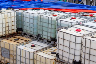 Paletlerin üzerine yığılmış orta büyüklükteki konteynırların (IBC 'lerin) yer aldığı yakın plan bir çekim. Sıvıların taşınması ve depolanması için kullanılan bu konteynırlar endüstriyel