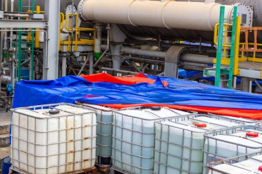 Bu resim birden fazla konteynırı olan, muhtemelen sıvı veya kimyasalları barındıran, açık havada depolanmış ve muşambayla kaplı bir endüstriyel tesisi tasvir ediyor. Sahnede çeşitli borular, endüstriyel
