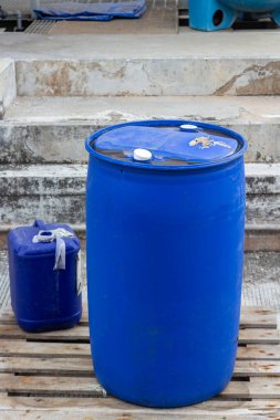 Bu görüntü açık havada duran mavi bir fıçı ve daha küçük mavi bir konteynırı gösteriyor. Varil, ahşap bir palete yerleştirilmiş iki taneden daha büyüktür. Her iki madde de endüstriyel bir ortam olduğunu gösteriyor.