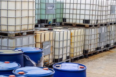 Görüntü, sanayi bölgesinde çeşitli mavi varilleri ve orta büyüklükteki konteynırları (IBC 'leri) gösteriyor. Bu kaplar genellikle sıvıların depolanması ve taşınması için kullanılır.