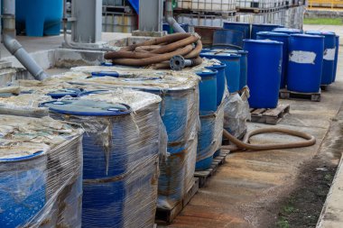 Mavi endüstriyel varillerin olduğu bir depo sahnesi, bazıları plastiğe sarılı, paletlere düzgünce yerleştirilmiş kahverengi bir hortumun yanında. Ayar, depolama, ulaşım ve