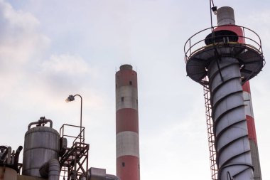 Bu görüntü endüstriyel bitki bacalarını bulutlu bir gökyüzüne uzanıp çevresel kirliliği sembolize ediyor. Sahne endüstriyel mimariyi, fabrikaların önemini ve etkilerini gözler önüne seriyor.