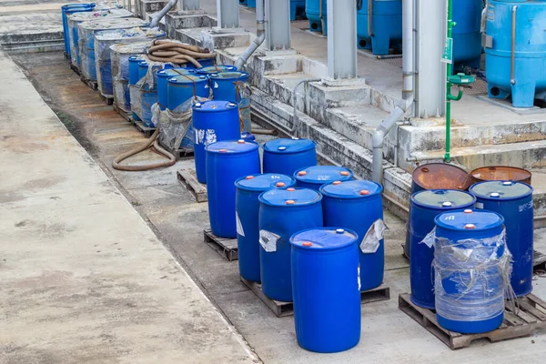 Resim, paletlerin üzerinde sayısız mavi plastik ve paslı metal varillerin bulunduğu bir endüstriyel depolama alanını gösteriyor. Varillerde kimyasallar ya da atıklar var gibi görünüyor, bir endüstri sahnesini vurguluyorlar ve