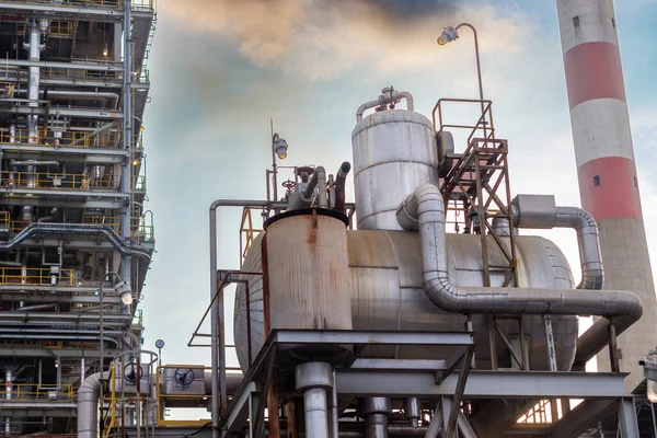 Bu resim, bulutlu bir gökyüzüne karşı boru, tank ve uzun bir baca ağının yer aldığı endüstriyel bir kompleksin ayrıntılı bir görüntüsünü sergiliyor. Karmaşık altyapı ölçeği vurguluyor ve...