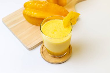 Canlı ve ferahlatıcı bir yıldız meyve püresi, ahşap bir altlığın üzerinde bir bardakta sunuluyor, yanında da tahtadan yapılmış taze yıldız meyveleri var. Bu görüntü sağlıklı beslenme ve tropikal yaşam hissi uyandırıyor.