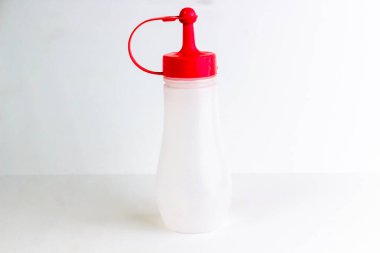 Parlak kırmızı kapaklı şeffaf bir sıkma şişe temiz beyaz bir yüzeye oturmuş, minimalist ve modern bir estetik yaratıyor, işlevselliği ve tasarım basitliğini vurguluyor..