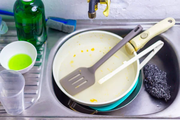 Kirli bir tava, mutfak gereçleri ve bulaşık deterjanı içeren tıkanmış bir lavabo, bir ev mutfağında yemek hazırlığından sonra günlük ev yüklerini yansıtıyor, sıradan ev işleri ve