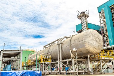 Endüstriyel rafineri kompleksi parlak bir gökyüzü altında karmaşık boru tesisatı ve işleme tankları gösteriyor.