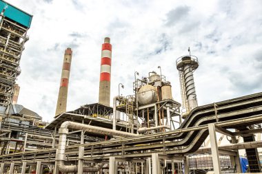 Sanayi manzarası bulutlu gökyüzüne karşı borular ve duman yığınlarıyla bir rafineri sergiliyor