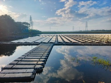 Bu resim, sakin bir göl üzerindeki yüzen bir güneş enerjisi santralini gözler önüne seriyor. Gökyüzünü ve bulutları incelikle yansıtıyor. Güneş panellerinin su üzerindeki yenilikçi kullanımı sürdürülebilir ve verimli