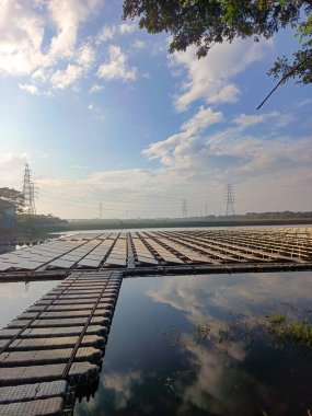 Durgun bir su kütlesi üzerinde yüzen geniş bir dizi güneş panelinin yer aldığı sakin bir manzara, pitoresk mavi gökyüzünü ve tüylü beyaz bulutları yansıtıyor. Bu yenilikçi yenilenebilir enerji sistemi