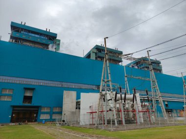 Uzun mavi binalar ve elektrikli trafoya sahip çitlerle çevrili bir bölgenin yer aldığı büyük bir elektrik santralinin alçak açılı çekimi. Tepedeki elektrik hatları gökyüzü boyunca uzanıyor ve üretilmiş olanları iletiyor.