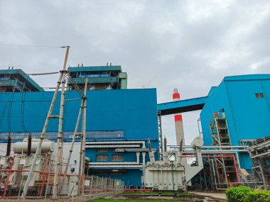 Düşük bir açıdan çekilen görüntüde kömür yakıtlı bir enerji santrali, mavi binaları bulutlu gökyüzü ile çelişiyor. Elektrikli trafolar ön planda duruyor.