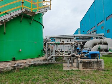 Erişim merdivenleri, karmaşık bir boru sistemi, vanalar ve pompalarla birlikte geniş yeşil bir depolama tankının bulunduğu bir sanayi tesisi. Tesiste şu karmaşıklıklar sergileniyor: