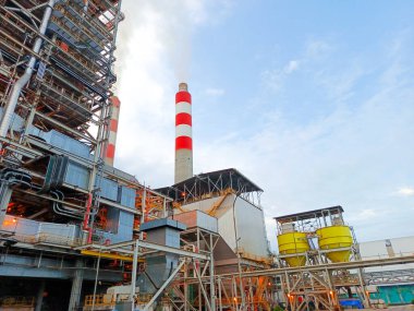 Düşük açılı bir çekimde geniş bir endüstriyel kompleks sergileniyor. Birden fazla bacası olan gökyüzü bulutlu. Görüntü bir enerji santralinin ölçeğini yakalıyor, karmaşık ağı vurguluyor