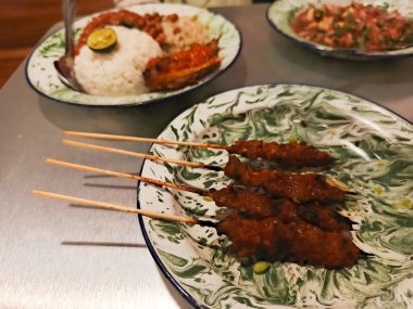 Bu resim Endonezya mutfağının enfes bir yayılımını sergiliyor, güzel desenli bir tabakta servis edilen satay şişleri, yanında pilav ve çeşitli Nasi lemak