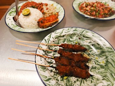 Ağız sulandıran Endonezya mutfağı lezzetleri, pirinç ve diğer geleneksel yemeklerle birlikte antika bir tabakta sunulan satay şişleri. Canlı renkler ve karmaşık kaplamalar.