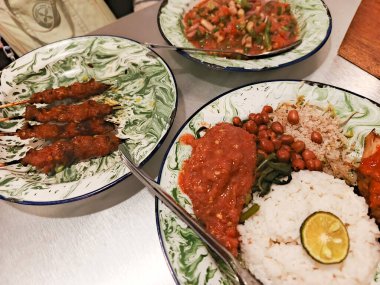 Sate Lilit ve Nasi Campur 'un da dahil olduğu Endonezya lezzetlerinin güzel desenli tabaklarda servis edildiği canlı bir görüntü. Yemekler ustalıkla düzenlenir, zenginlerin altını çizer.