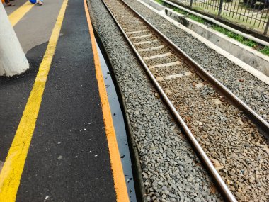 Zemin seviyesindeki bir perspektif, tren istasyonunun kenarındaki rayların yanında sarı bir güvenlik hattı barındıran bir perspektifi yakalıyor. Sahne toplu taşımanın altyapısını vurguluyor