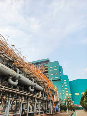 Düşük açılı çekimde genişleyen bir sanayi fabrikası, karmaşık boru ve yapısal elementlerden oluşan ağını vurguluyor. Bitkinin berrak mavi gökyüzüne karşı yan yana gelmesi...