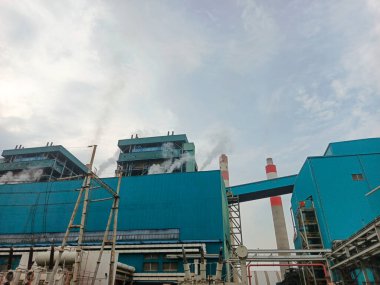 Gökyüzüne buhar yayan, birden fazla yapıya, duman yığınlarına ve borulara sahip büyük bir enerji santralinin görüntüsü. Sanayi kompleksi enerji için gerekli makine ve altyapıyı sergiliyor