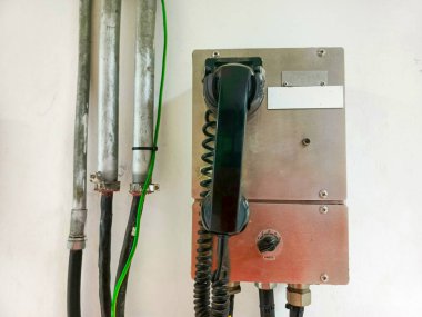 Yanında metal borular olan beyaz bir duvara monte edilmiş endüstriyel bir telefonun yakın plan çekimi. Metalik bir kutunun içine gizlenmiş olan telefon, sarmalanmış bir kablo ve bir cep telefonu içeriyor. Kablolar ve