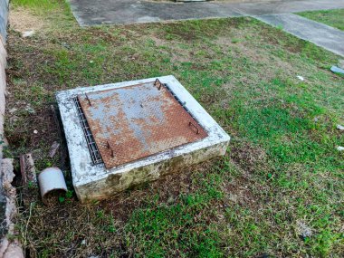 Yüksek açılı bir çekim, betonla kaplanmış ve çimlerle çevrili paslı bir rögar kapağını tasvir ediyor. Beton yapı ve metal kapak, uzun süre kullanıldığını gösteren yıpranma ve yaşlanma belirtileri gösteriyor.