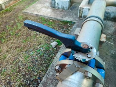 Yakın plan çekimde, beton ve çimen elementleri bulunan endüstriyel metal bir boru ve levyeli bir vana sergileniyor. Kapakçık ve boru karmaşık bir şekilde birbirine bağlı, metal bir flanş, vidalar,