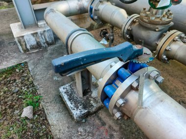 Açık havada endüstriyel boru hattı sisteminin bir bölümünü gösteren yakın çekim. Görüntü, metal borular, valfler ve flanşlar gibi kompleksi vurgulayarak birbirine tutturulmuş çeşitli bileşenleri içerir.