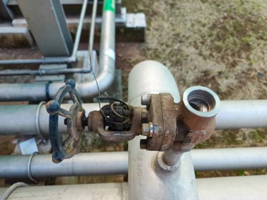 Bu resim, belirgin bir kapakçık ve karmaşık bir metal boru düzeneği içeren endüstriyel bir boru sisteminin ayrıntılı bir yakın çekimini sergiliyor. Dış duvarın yakınındaki dış mekan gölgeleri...