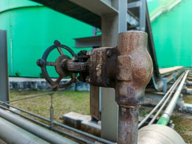 Yakın plan çekimde, karmaşık bir boru hattı sistemine aşırı derecede aşınmış bir endüstriyel kapak sergileniyor. Kapakçık önemli bir pas ve oksitlenme gösteriyor. Yeşil depolama tankları arkaplan oluşturur,