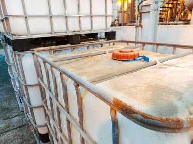 Metal çerçeveli bir sanayi konteynırı yığını, toplu sıvılarla başa çıkmak için kullanılan lojistik altyapıyı gösteriyor. Konteynırlar açık hava depolama ve endüstri kullanımından eskimiş bir estetik sergiliyor.