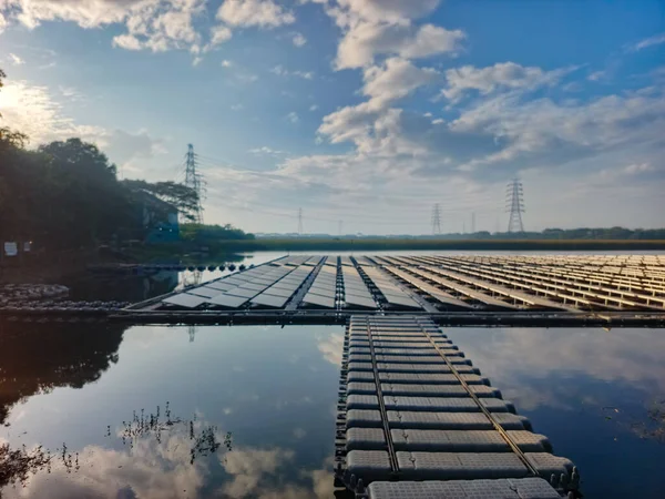 Yakaladığımız, sakin bir gölde yüzen bir güneş paneli çiftliğinin geniş açılı görüntüsü. Paneller, gökyüzünü ve etrafındaki ağaçları yansıtan mesafeye kadar uzanıyor. Panellerden oluşan bir yürüyüş yolu gözü açar.