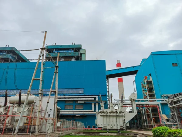 Düşük bir açıdan çekilen görüntüde kömür yakıtlı bir enerji santrali, mavi binaları bulutlu gökyüzü ile çelişiyor. Elektrikli trafolar ön planda duruyor.