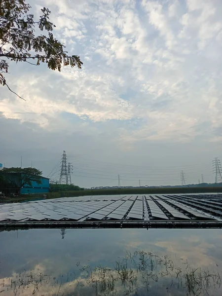 Güneş çiftliğinin büyüleyici bir görüntüsü zarif bir şekilde suyun üzerinde yüzüyor, yukarıdaki parçalı bulutlu gökyüzünü yansıtıyor. Sahne, elektrik hatları ve yemyeşil bir zemin üzerine kurulmuş.