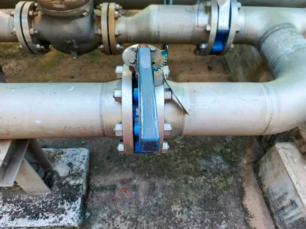 Kapakçık ve flanj bağlantıları olan endüstriyel bir boru sistemini gösteriyor. Gümüş renkli borular ve mavi vananın sapı endüstriyel ortamda bir tesisat sistemi olduğunu gösteriyor.,