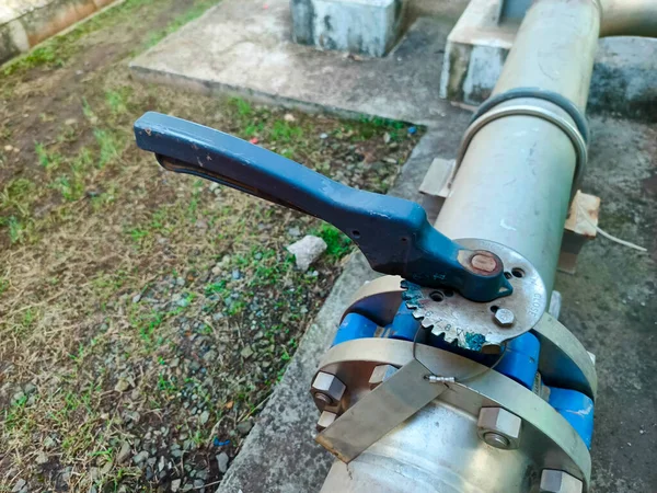 Yakın plan çekimde, beton ve çimen elementleri bulunan endüstriyel metal bir boru ve levyeli bir vana sergileniyor. Kapakçık ve boru karmaşık bir şekilde birbirine bağlı, metal bir flanş, vidalar,