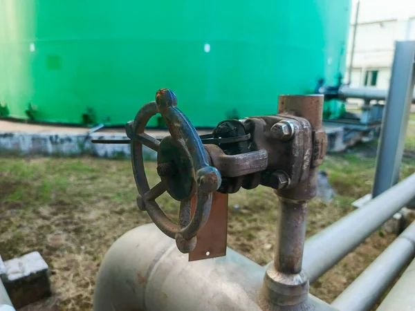 Yakın çekim, eski, paslı bir endüstriyel vananın eskimiş dokusunu büyük bir yeşil depolama tankının arka planına yapıştırır. Boru hattı, aşınma ve oksitlenme izleri gösteriyor.,
