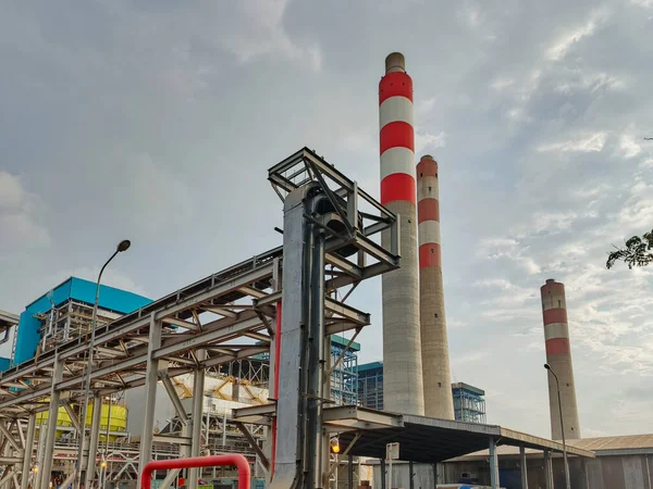 Önde gelen kırmızı ve beyaz çizgili bacaları olan bir sanayi enerji santralini ele geçiren zemin seviyesinde bir perspektif. Komplekste metal yapılar, borular ve altyapı setleri yer alıyor.